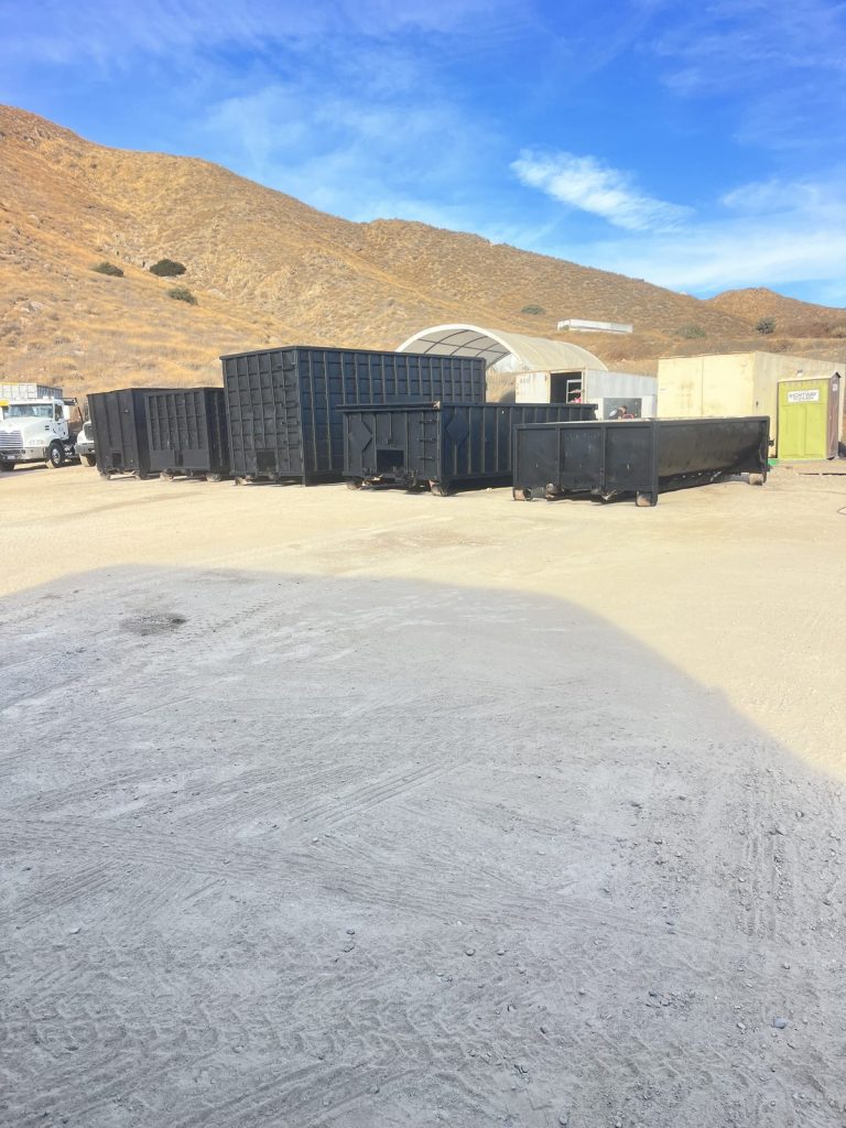 Several empty black roll-off dumpsters lined up on a paved yard below brown hills near Corona, CA