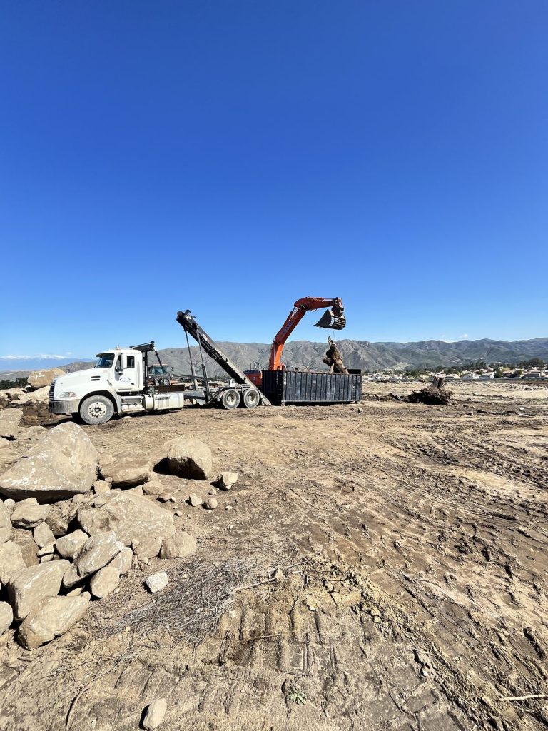 Orange excavator loading dirt and rock into a black roll-off dumpster beside a JD Transport truck on a hillside grading site