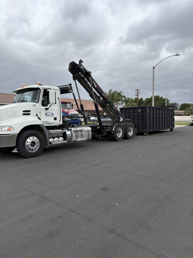 JD Transport Mack roll-off truck unloading a large black roll-off dumpster curbside on a residential street in Riverside, CA