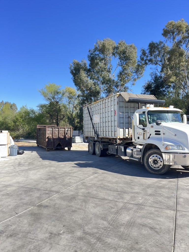 White JD Transport Mack roll-off truck carrying a tall high-side roll-off container across a paved lot on a sunny day