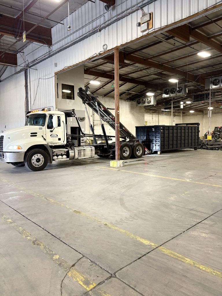 White JD Transport Mack roll-off truck with hoist raised inside a covered service bay with a black roll-off container in Riverside, CA