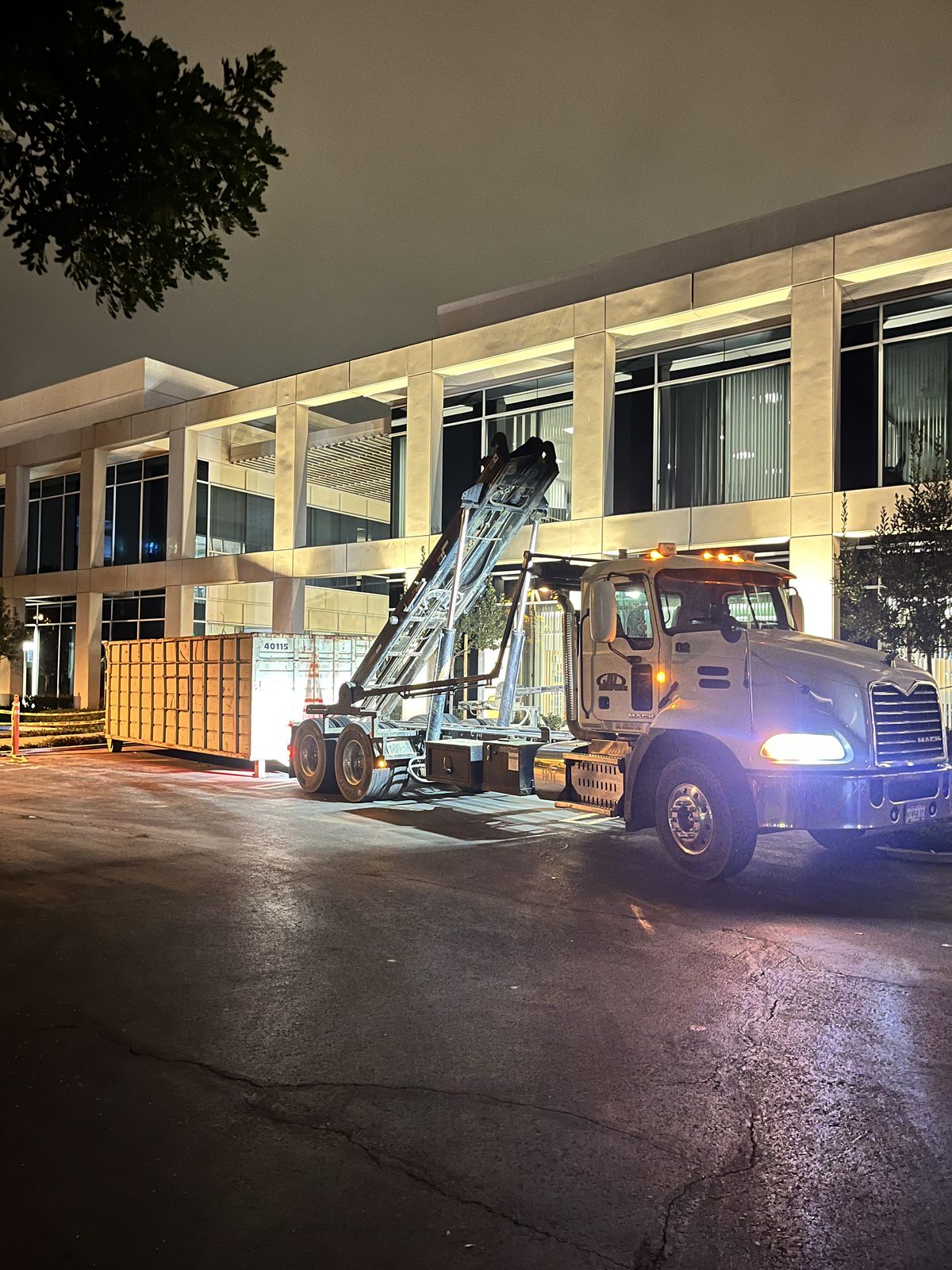 JD Transport roll-off truck with headlights on and hoist raised, swapping a yellow roll-off container outside a lit office building at night
