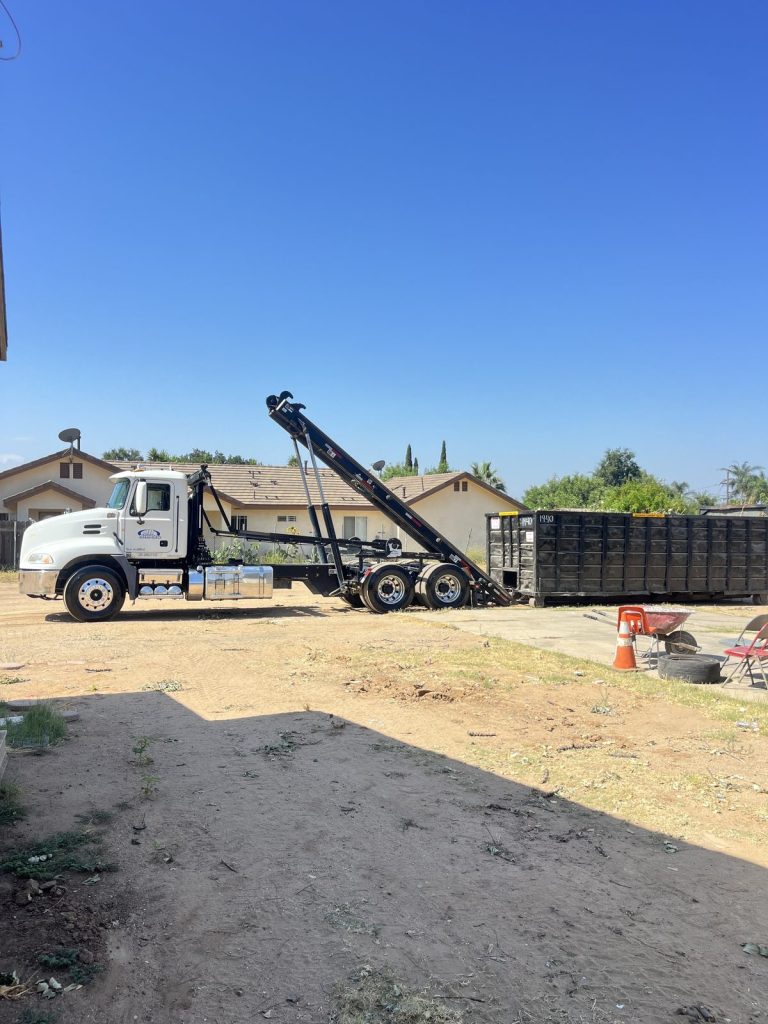 JD Transport roll-off truck beside a freshly delivered black roll-off dumpster on a dirt residential lot next to homes