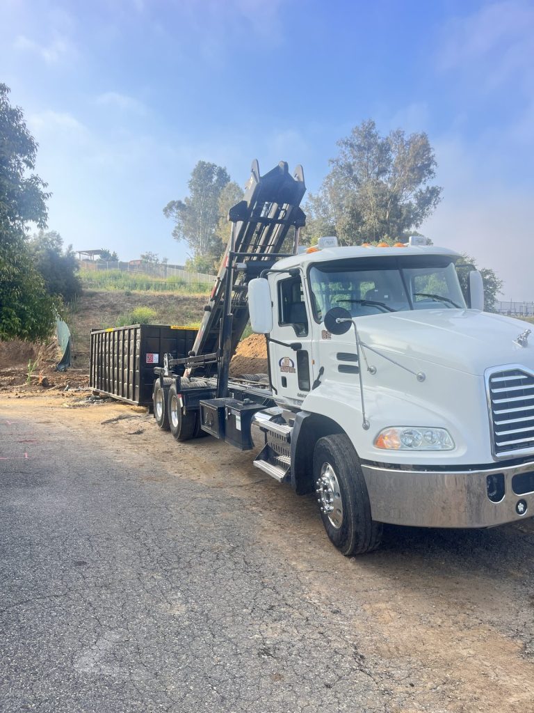 White JD Transport roll-off truck with hoist raised setting a black roll-off dumpster on a dirt shoulder at an early-morning jobsite