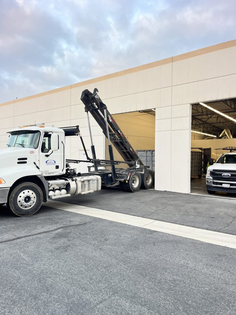JD Transport roll-off truck with hoist raised beside an open warehouse loading bay at an Inland Empire distribution facility