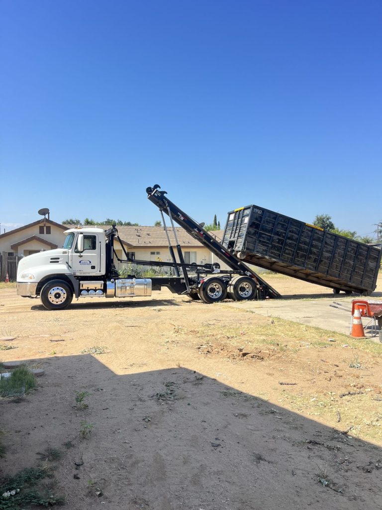 JD Transport roll-off truck with hoist raised, sliding a black roll-off dumpster onto a dirt residential lot