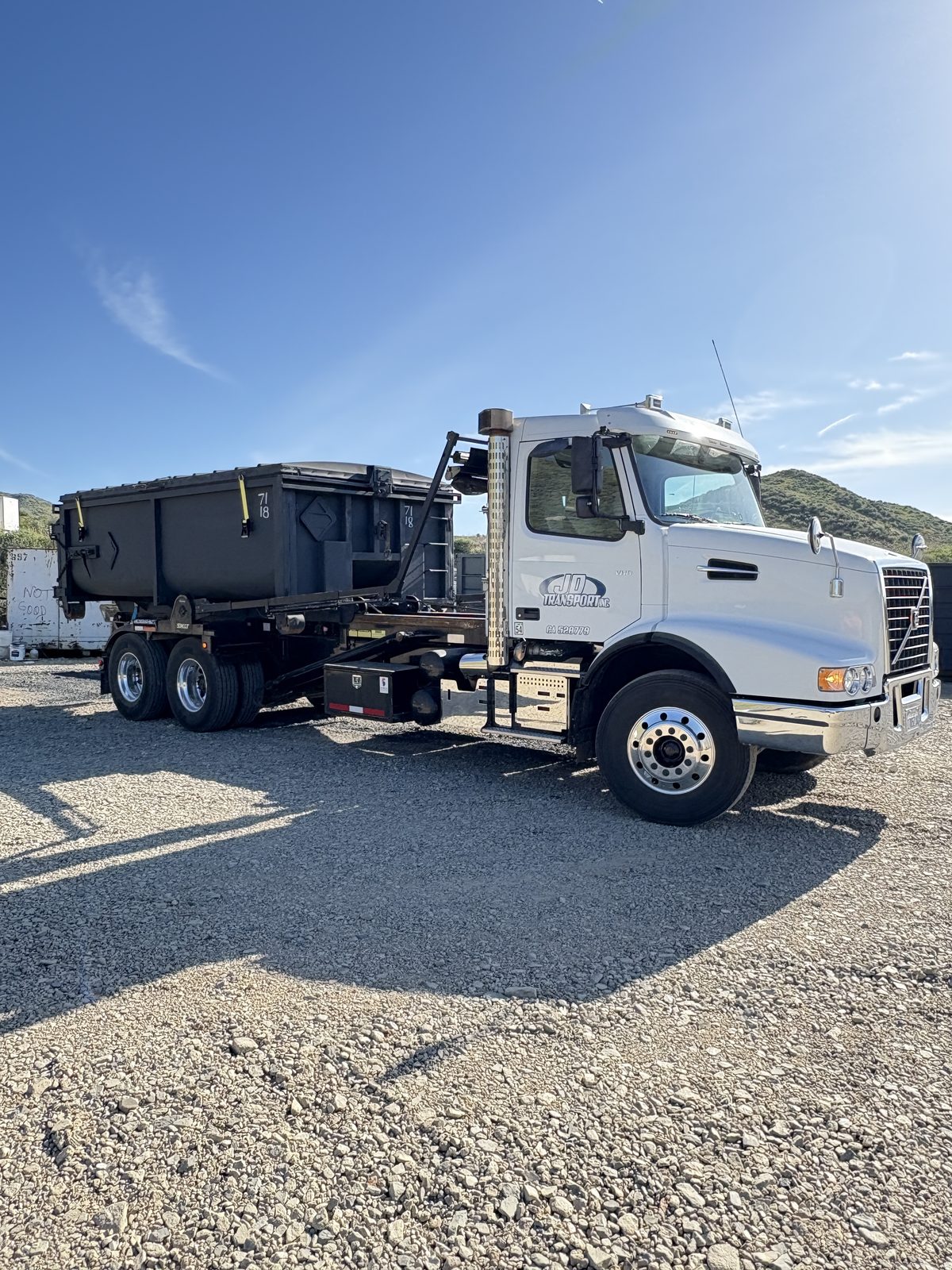 White JD Transport roll-off truck carrying a black roll-off dumpster across a gravel yard near Corona, CA