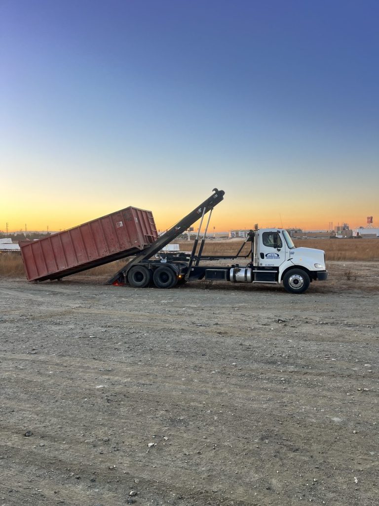JD Transport roll-off truck with hoist raised, loading a red roll-off container on an open dirt jobsite at sunrise