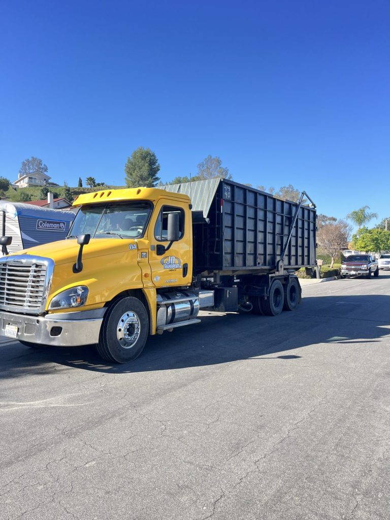 Yellow JD Transport Freightliner roll-off truck hauling a full roll-off dumpster down a residential street on a clear day